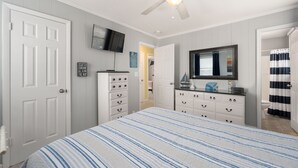 Ein Schlafzimmer mit einem Bett, einer Kommode, einem Schminktisch mit Spiegel, einem Deckenventilator und einem an der Wand befestigten Fernseher.