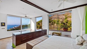 A modern bedroom with a large bed, a vanity with a mirror, and a view of a coastal landscape through large windows. - Suite 2 - Casa Samo Conchas Chinas (Puerto Vallarta)