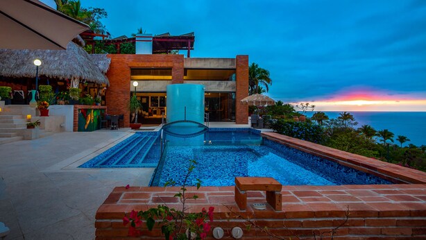 A resort pool with a view of the ocean at sunset, a thatched-roof bar, and a modern building with a glass entrance. - Suite 2 - Casa Samo Conchas Chinas (Puerto Vallarta)