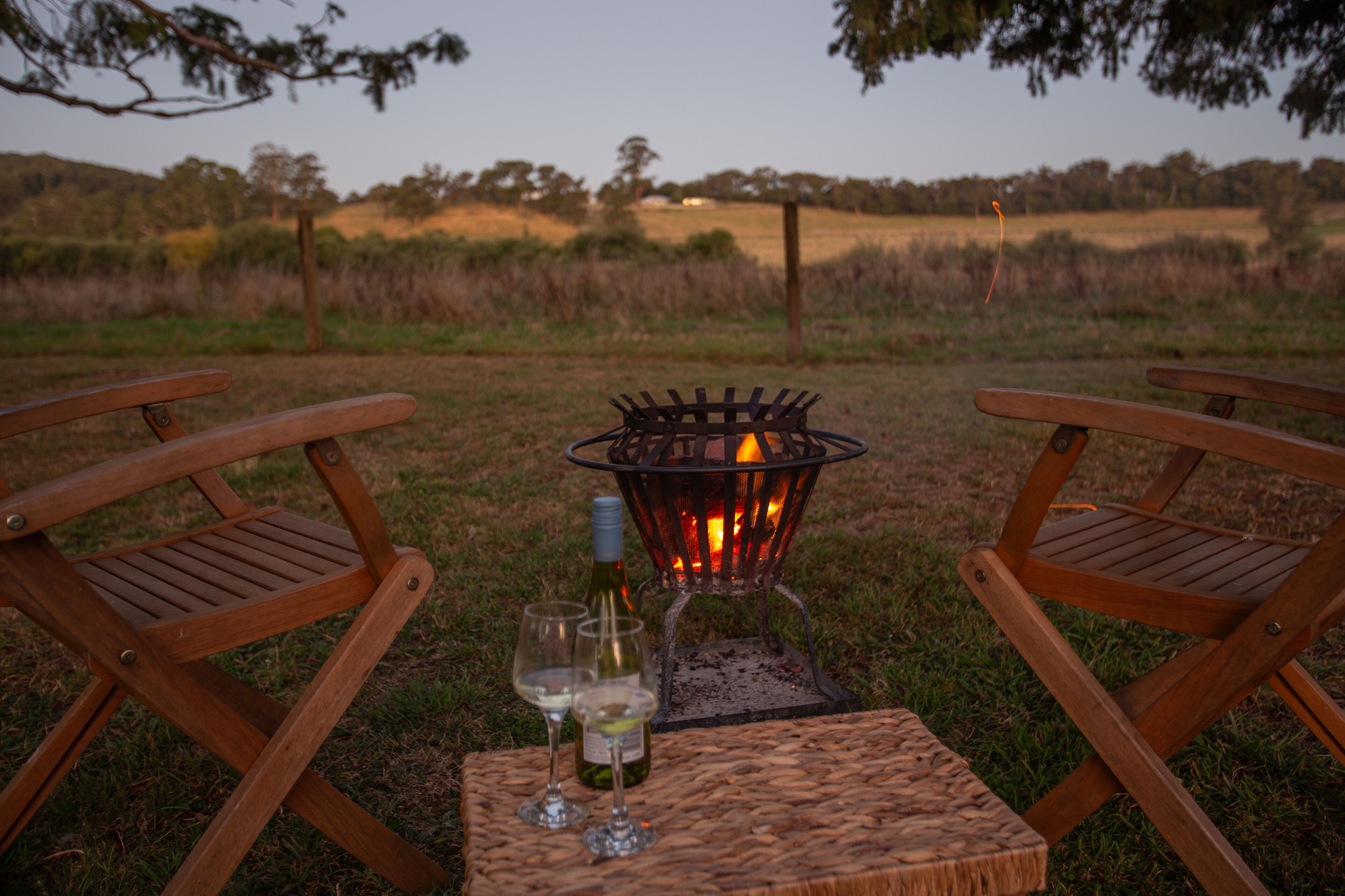 Two wooden chairs facing each other with a small table between them, a bottle of wine, and two wine glasses on the table. In the background, there is a fire pit with flames.