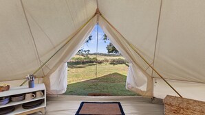 Een tent met uitzicht op een grasveld. Binnenin staat een bed met een matras, een plank met borden en een mand.