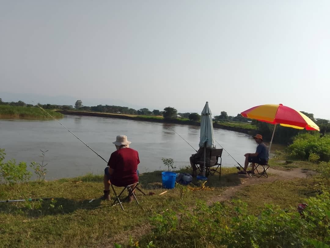 Two people fishing by a calm river with a red and yellow umbrella providing shade.