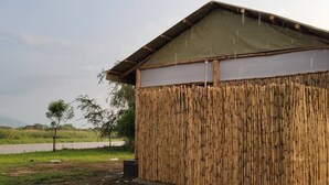 A bamboo structure with a slanted roof, likely a simple shelter or hut, situated in a rural setting with a road and vegetation in the background. - Semliki Fishing River Camp (Bweramule)