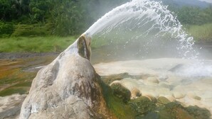 A geyser with water erupting into the air, surrounded by a natural landscape with hills and greenery. - Semliki Fishing River Camp (Bweramule)