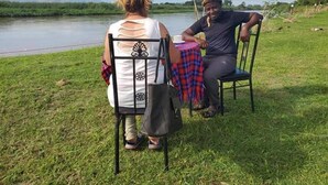 Two people seated at an outdoor table with a view of a river. - Semliki Fishing River Camp (Bweramule)