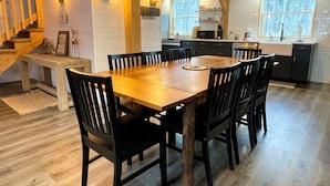 Una mesa de comedor de madera con sillas, una cocina con paredes de ladrillo blanco y una escalera con barandillas de madera.