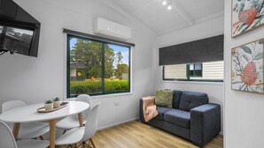 A modern living room with a grey sofa, white chairs, a round table, and a large window with a view of greenery. - Creswick Holiday Park (Creswick)