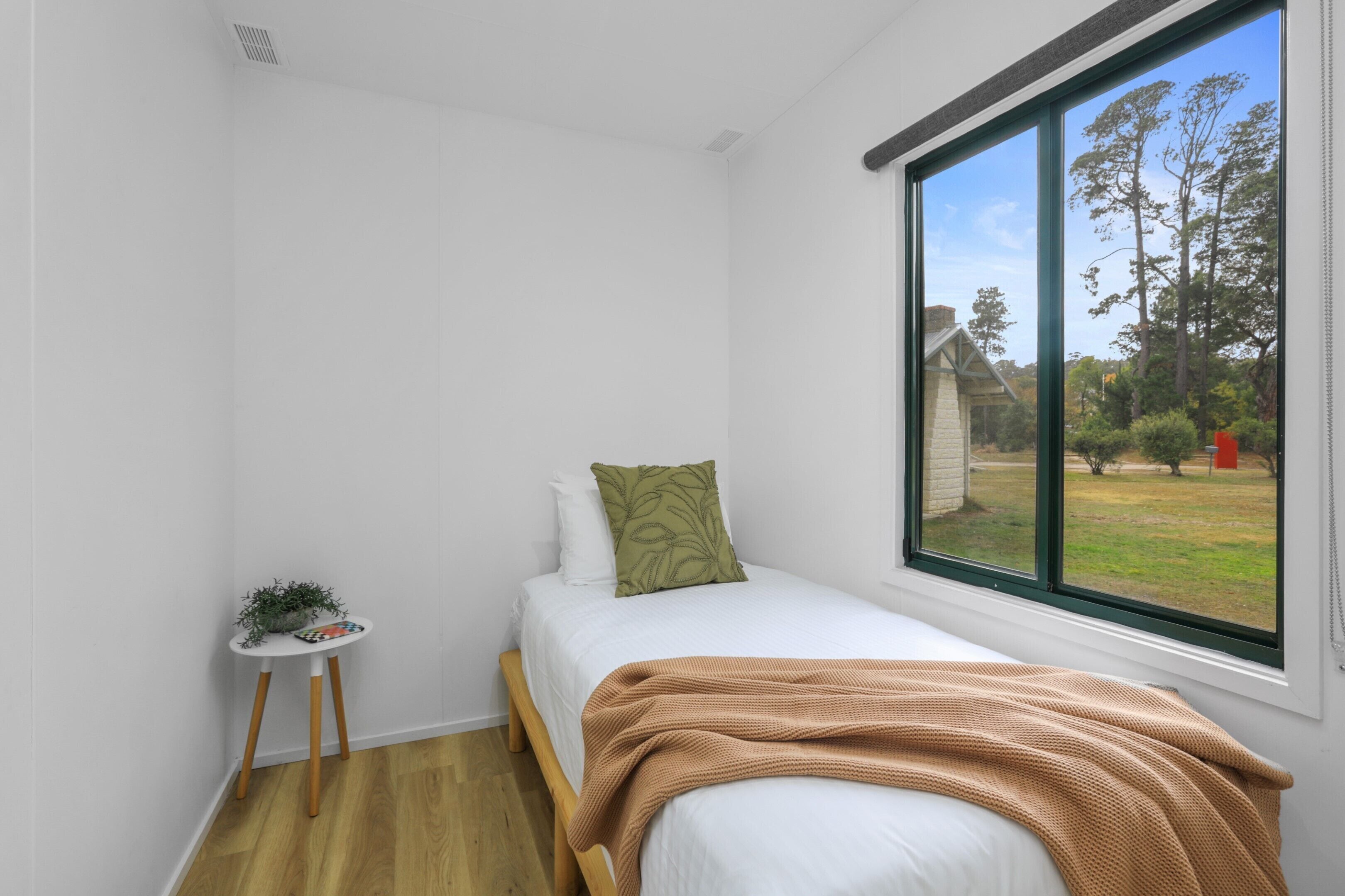 Ein Schlafzimmer mit einem Einzelbett, einem kleinen Nachttisch mit Pflanze und einem Fenster mit Blick ins Freie.