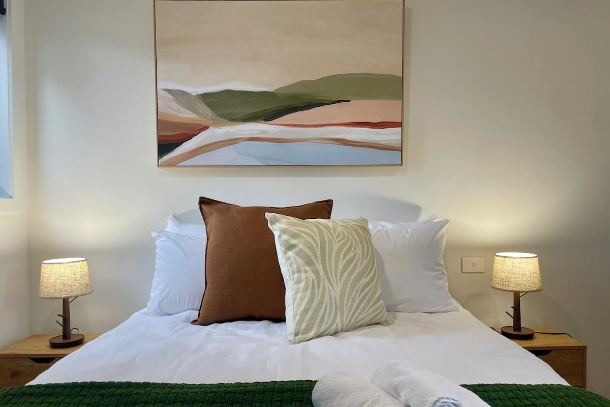 Una cama bien tendida con sábanas blancas, dos almohadas marrones y una blanca, una manta verde y dos lámparas de noche.