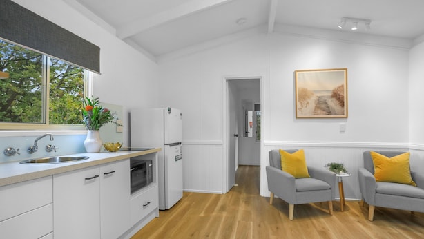 A modern kitchen with white cabinetry, a stainless steel sink, and a refrigerator. There is a window with a view of trees, and a painting on the wall. - Creswick Holiday Park (Creswick)