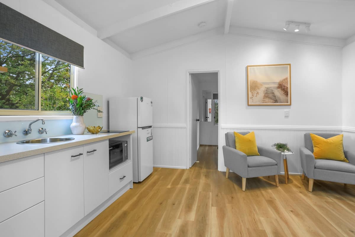 Una cocina moderna con armarios blancos, un lavamanos de acero inoxidable y un refrigerador. Hay una ventana con vistas a árboles y un cuadro en la pared.