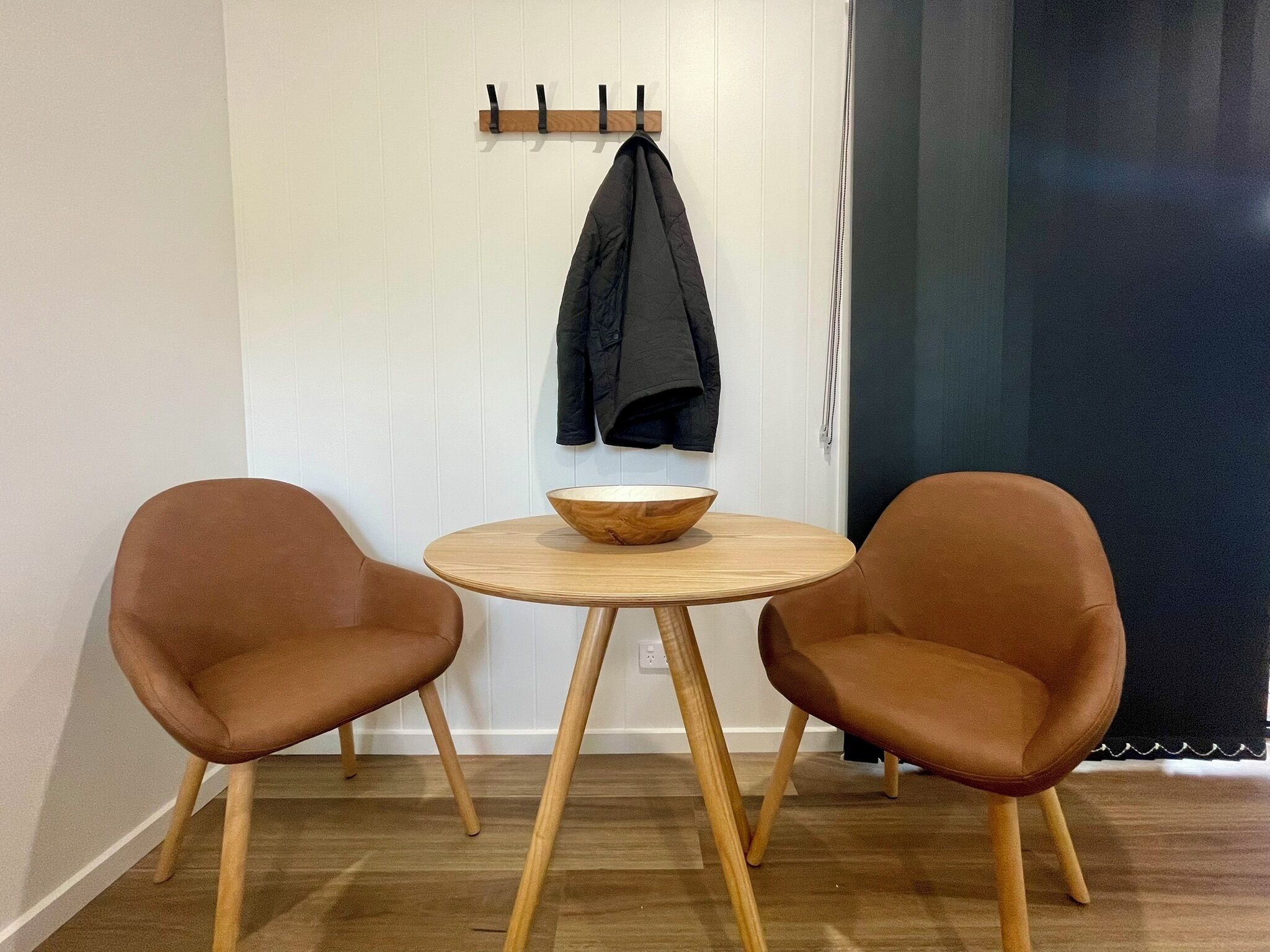 Una habitación con dos sillas marrones alrededor de una pequeña mesa de madera con un cuenco. Una chaqueta negra cuelga de un perchero de madera en la pared.