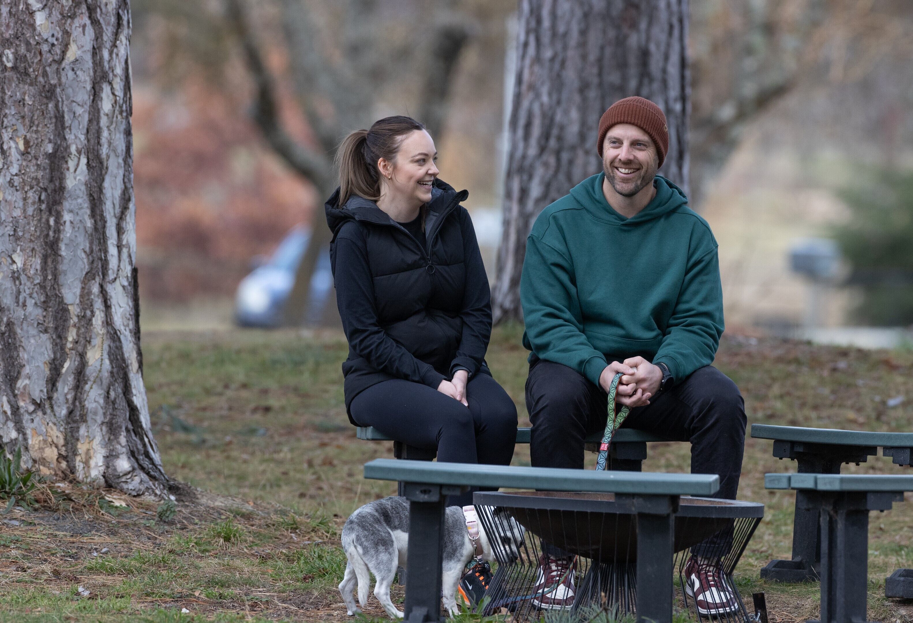 Two people sitting on a bench in a park with trees and a dog nearby.