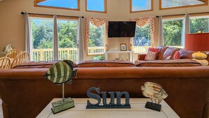 A living room with a brown sofa, a white coffee table, and a TV mounted on the wall. - Seas The Day - Oceanside 4 BR Home w/Private Pool - Short walk to shops/restaurants (Duck)