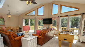 A spacious living room with a brown leather sofa, a white cabinet, a wooden foosball table, and a television mounted on the wall. - Seas The Day - Oceanside 4 BR Home w/Private Pool - Short walk to shops/restaurants (Duck)