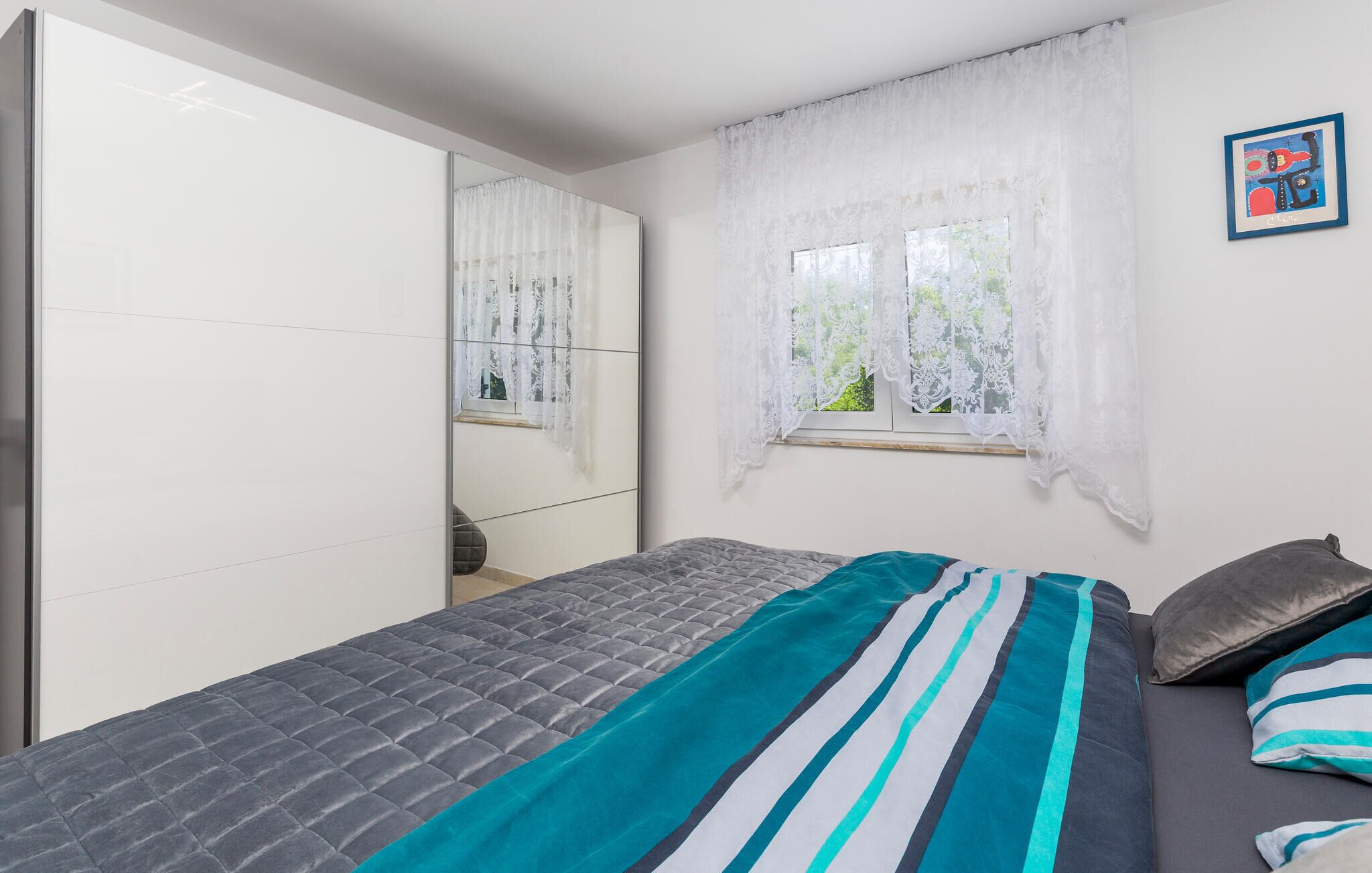 A bedroom with a bed, a wardrobe, a window with curtains, and a framed picture on the wall.