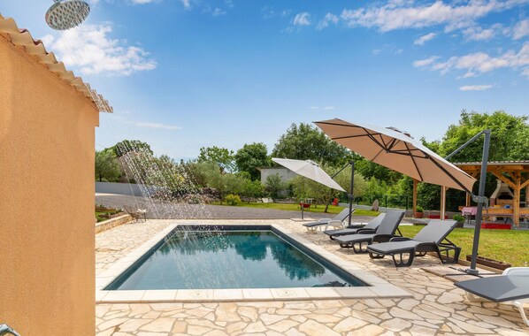 A swimming pool with a stone border, surrounded by lounge chairs and umbrellas, with a clear sky and greenery in the background.