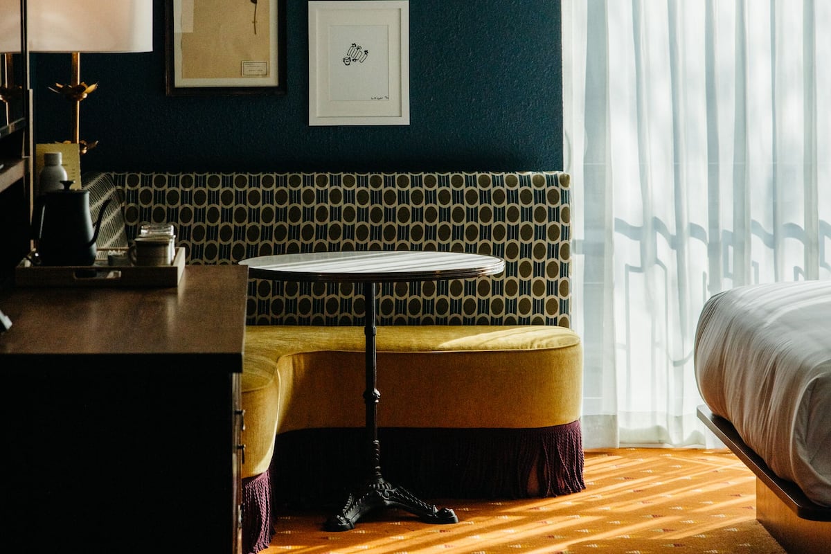 Een hotelkamer met een bed, een bureau met een lamp, een bijzettafeltje, een bankje en een behang met een patroon.