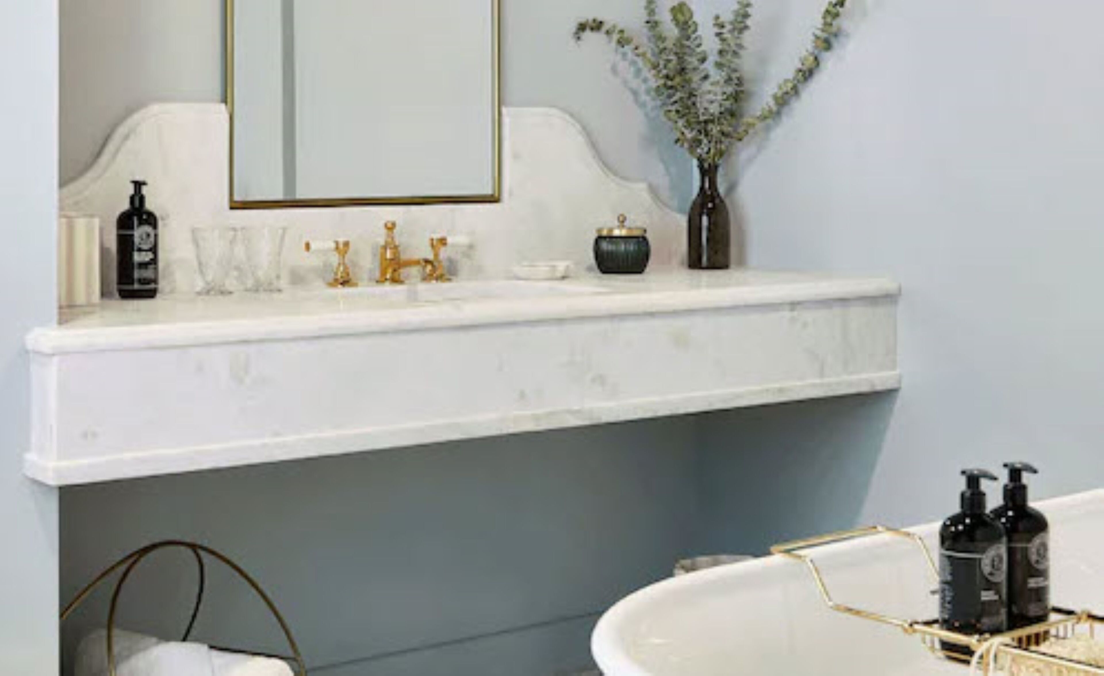 Une salle de bain avec un lavabo en marbre blanc, des robinets dorés, un vase avec des plantes vertes et deux bouteilles noires posées sur le comptoir.