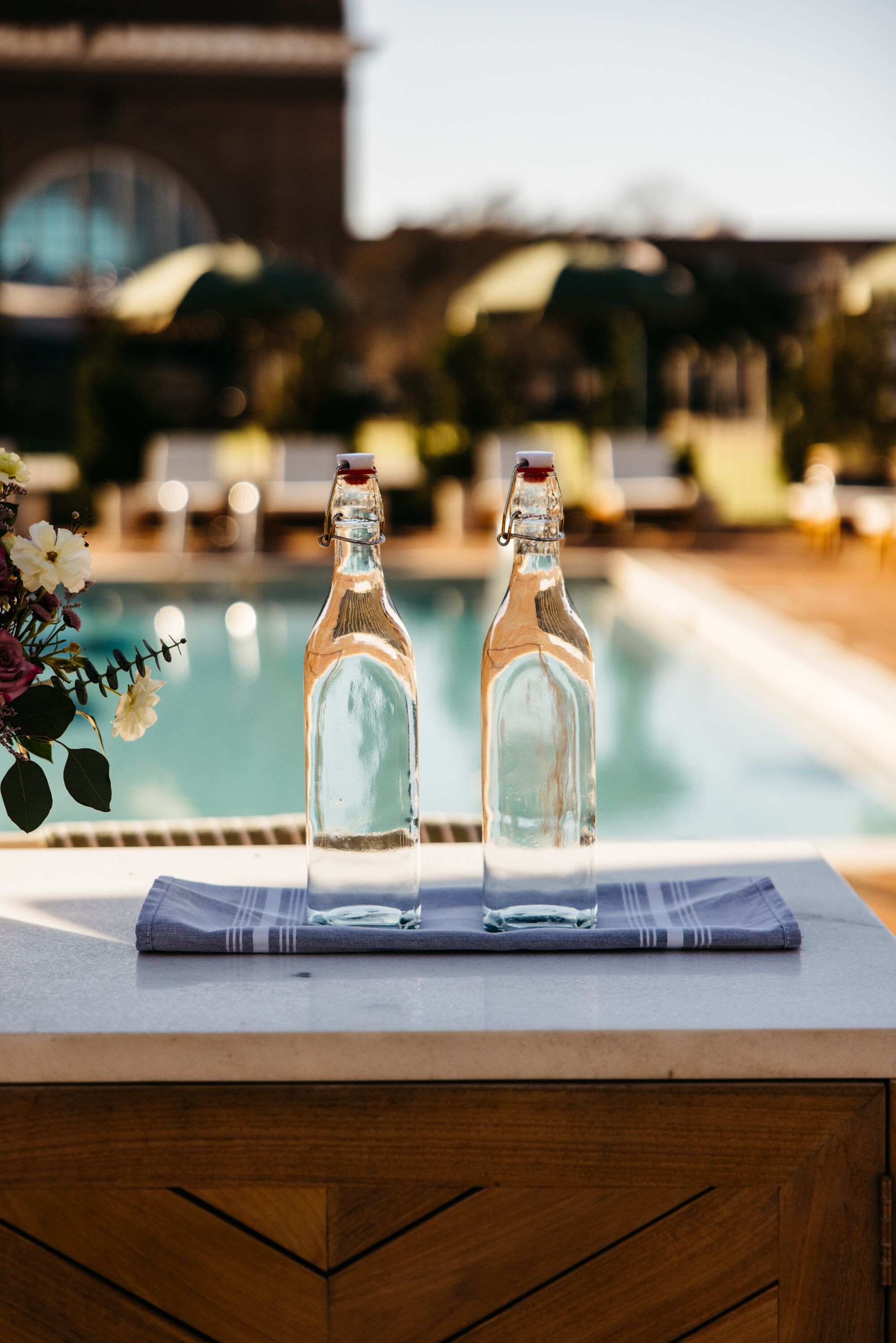 Two empty glass bottles on a table by a poolside with a wooden cabinet below.