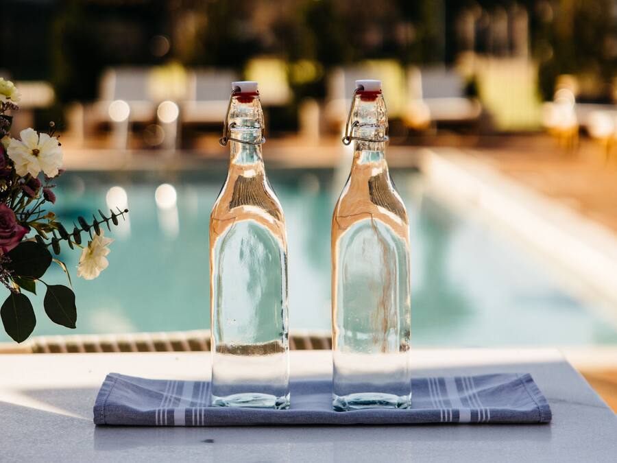 Two empty glass bottles on a table by a poolside with a wooden cabinet below.