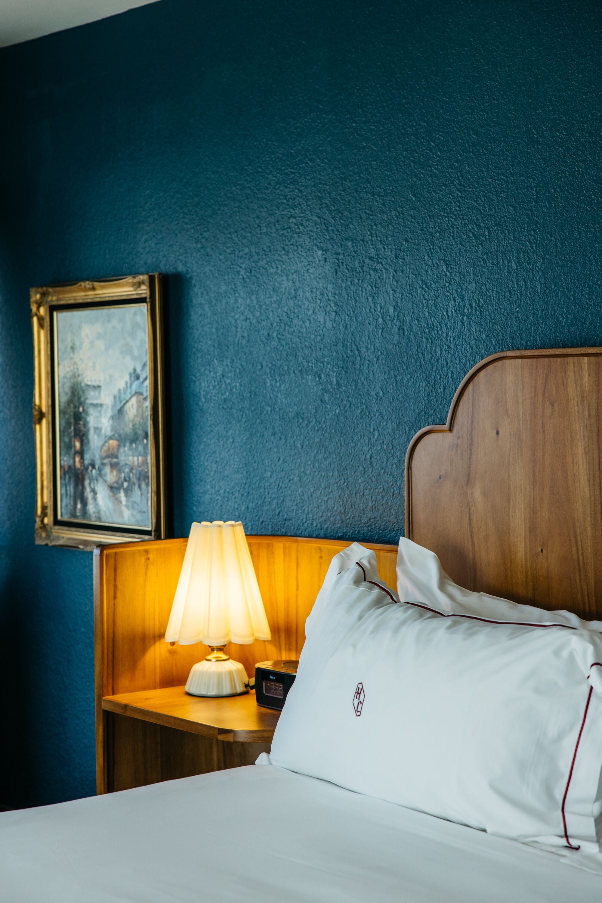 A hotel room with a wooden headboard, a bedside lamp, a framed picture on the wall, and a white pillow with red trim.