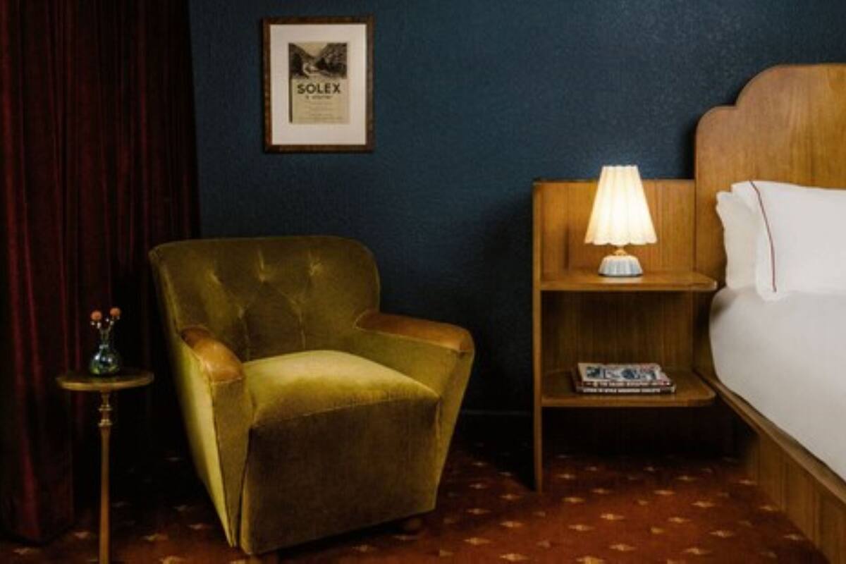 Een hotelkamer met een groene fauteuil, een houten nachtkastje met een lamp en een bed met witte lakens.