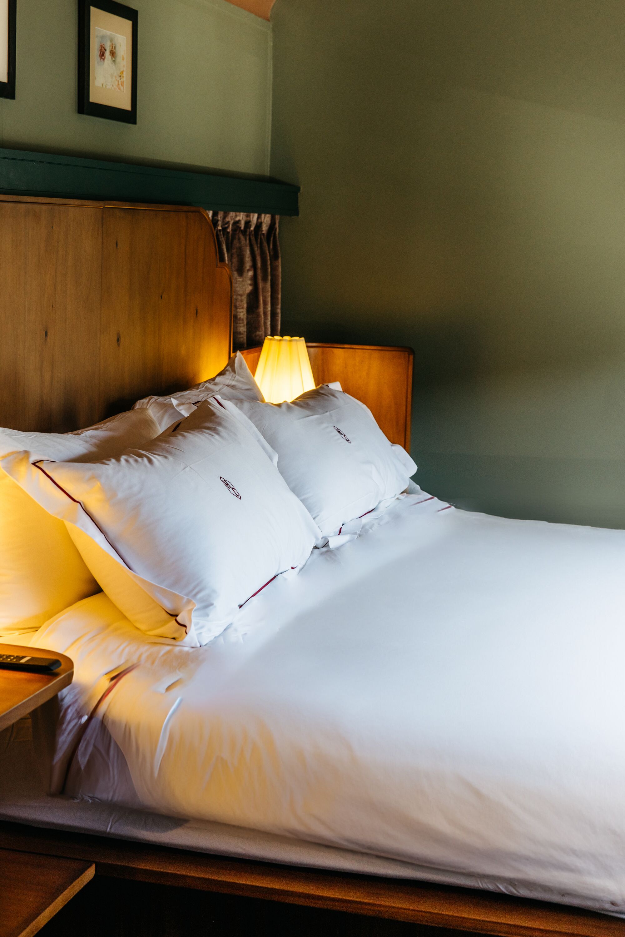 Una cama bien hecha con sábanas y almohadas blancas, un cabecero de madera y una lámpara de noche.