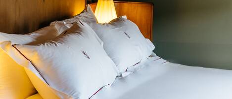 Una cama bien hecha con sábanas y almohadas blancas, un cabecero de madera y una lámpara de noche.