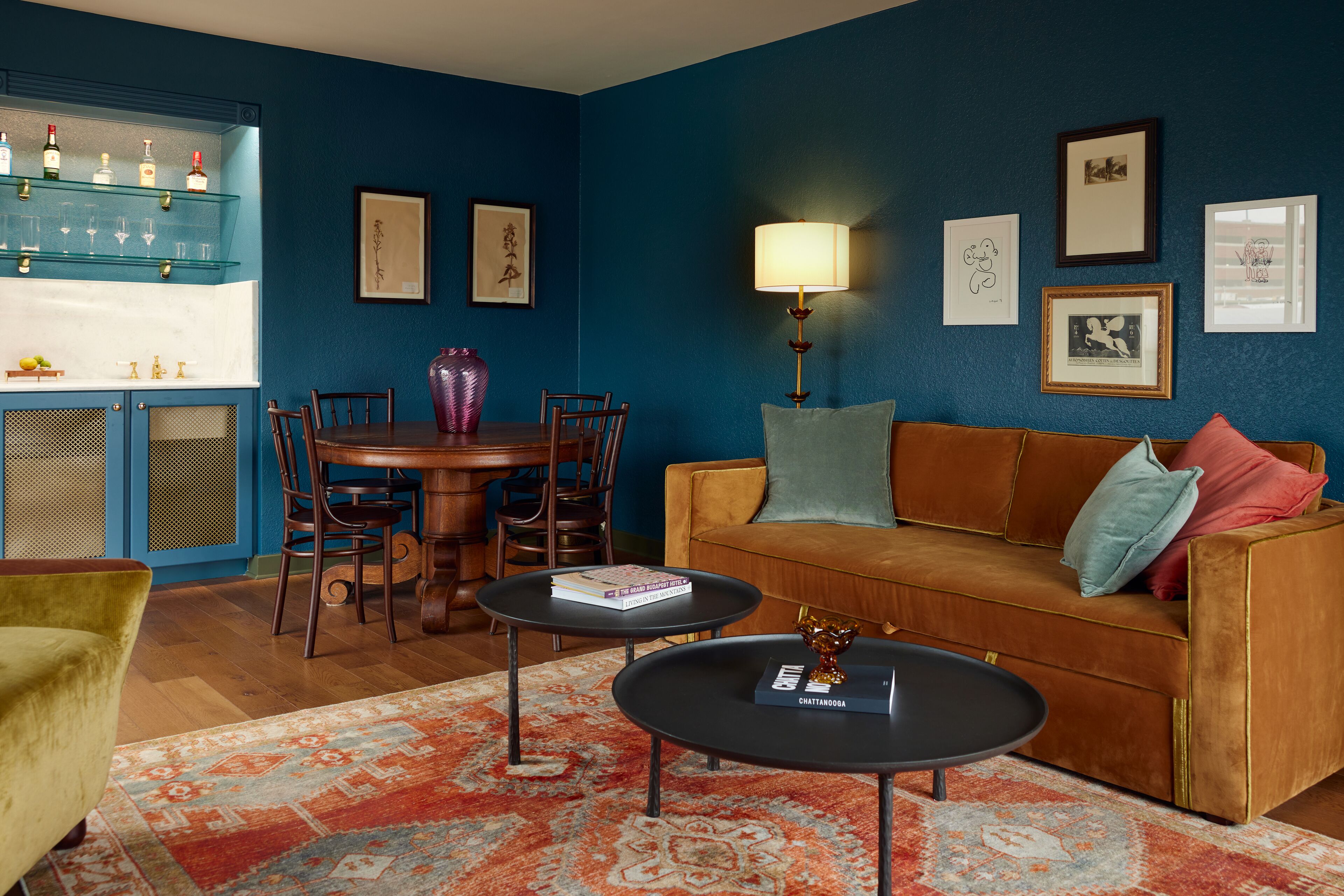 Un salon avec un mur bleu, un canapé marron, une table basse, une table à manger avec des chaises et un coin bar avec des étagères et des bouteilles.