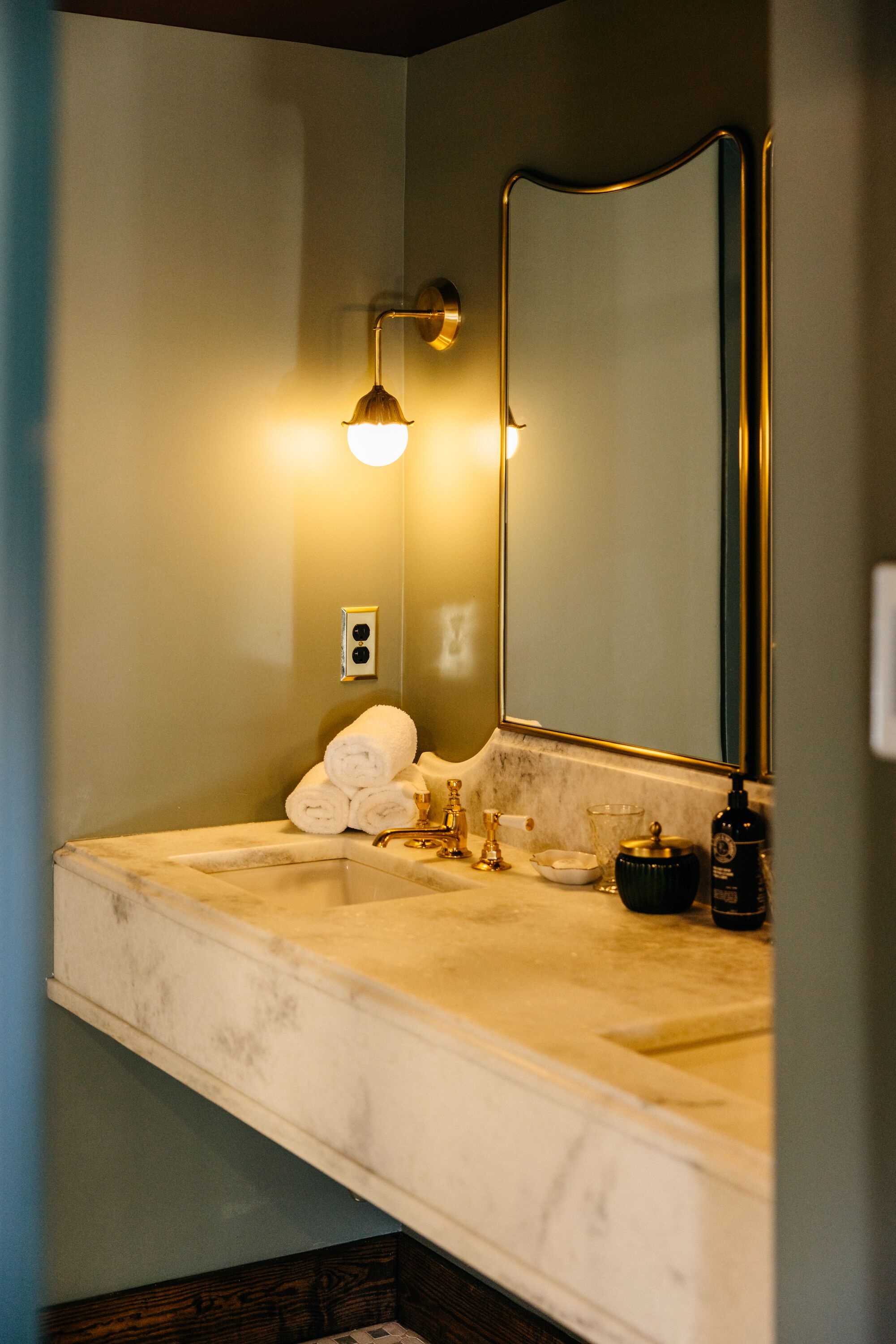 A bathroom with a marble sink, gold fixtures, a large mirror, and a wall-mounted light.