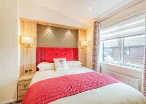 A hotel room with a red headboard, a large bed with white bedding, two bedside lamps, a window with blinds, and a view of the outside. - Sea View Lodge 3 Pet Friendly (Kirkcudbright)