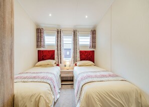 Una habitación con dos camas, cada una con cabecero rojo, una mesita de noche con lámpara y una ventana con cortinas.