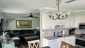 Ein Wohnzimmer mit einer Couch, einem Couchtisch und einem Esstisch mit Stühlen. Es gibt eine Küchenzeile mit weißen Schränken, einer Spüle und einem Herd. Ein Deckenventilator und eine Pendelleuchte sind zu sehen.