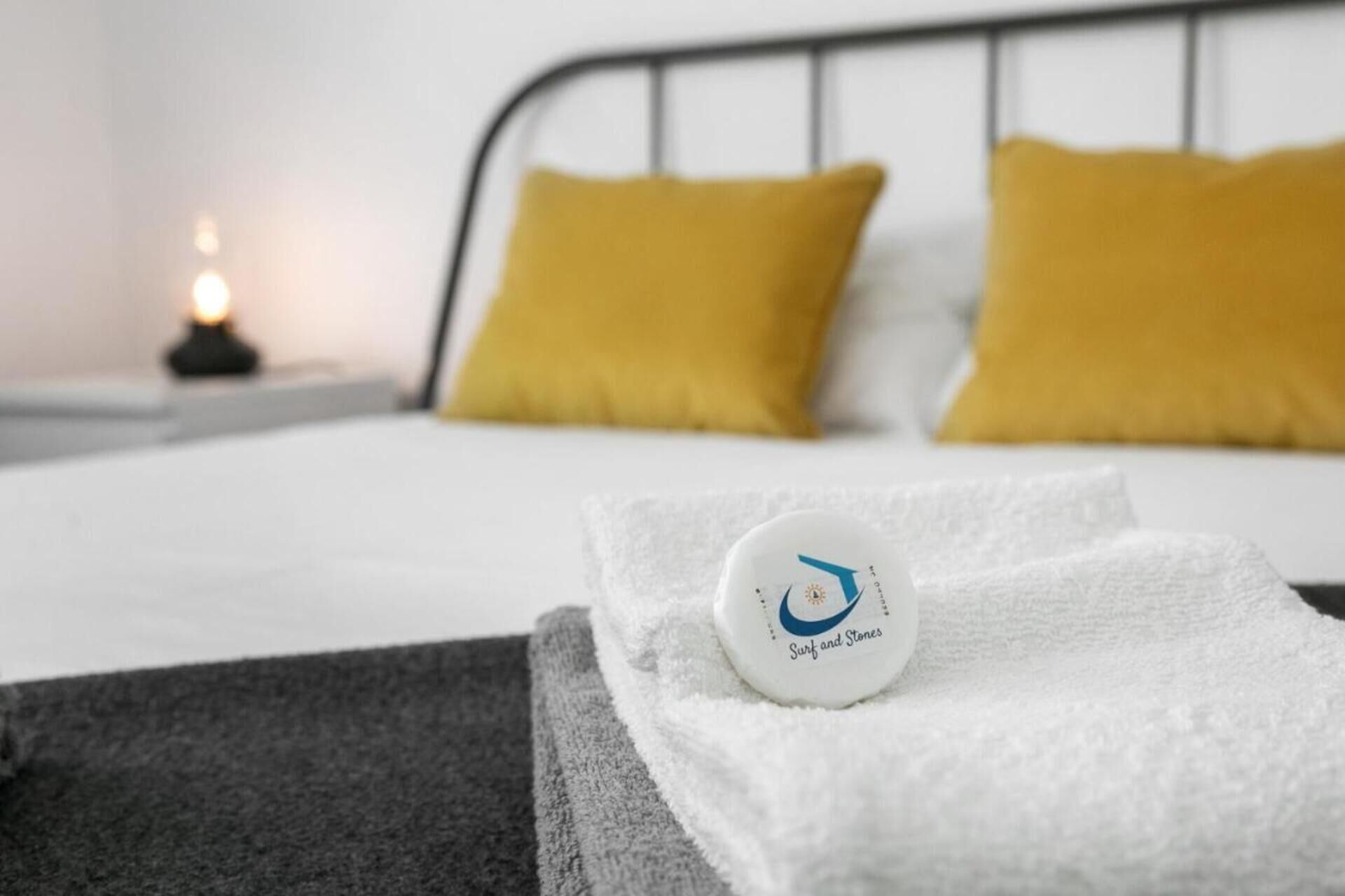 A neatly made bed with white bedding and two mustard yellow pillows, a white towel, and a round object with text on it.