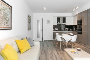 Une cuisine moderne avec des meubles blancs, un coin repas doté d’une table et de chaises blanches, et un canapé agrémenté de coussins jaunes.