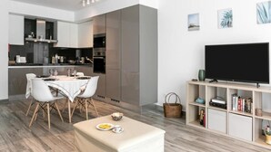 Une cuisine moderne avec des meubles blancs, un coin repas avec une table et des chaises blanches, et un téléviseur posé sur une étagère en bois.
