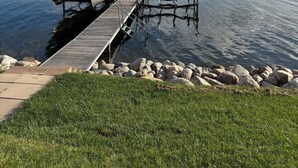 Un muelle que se adentra en aguas tranquilas, con un barco cubierto y sillas, rodeado de rocas y hierba.