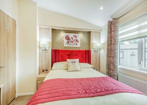 A bedroom with a large bed, a red headboard, two bedside lamps, a floral artwork above the bed, and a window with a view of a building outside.
