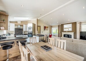 A modern kitchen with built-in appliances, a dining area with a wooden table and chairs, and a living area with a sofa and fireplace. - Country and modern cladded lodge. (Kirkcudbright)
