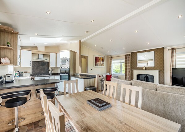A modern kitchen with built-in appliances, a dining area with a wooden table and chairs, and a living area with a sofa and fireplace. - Country and modern cladded lodge. (Kirkcudbright)