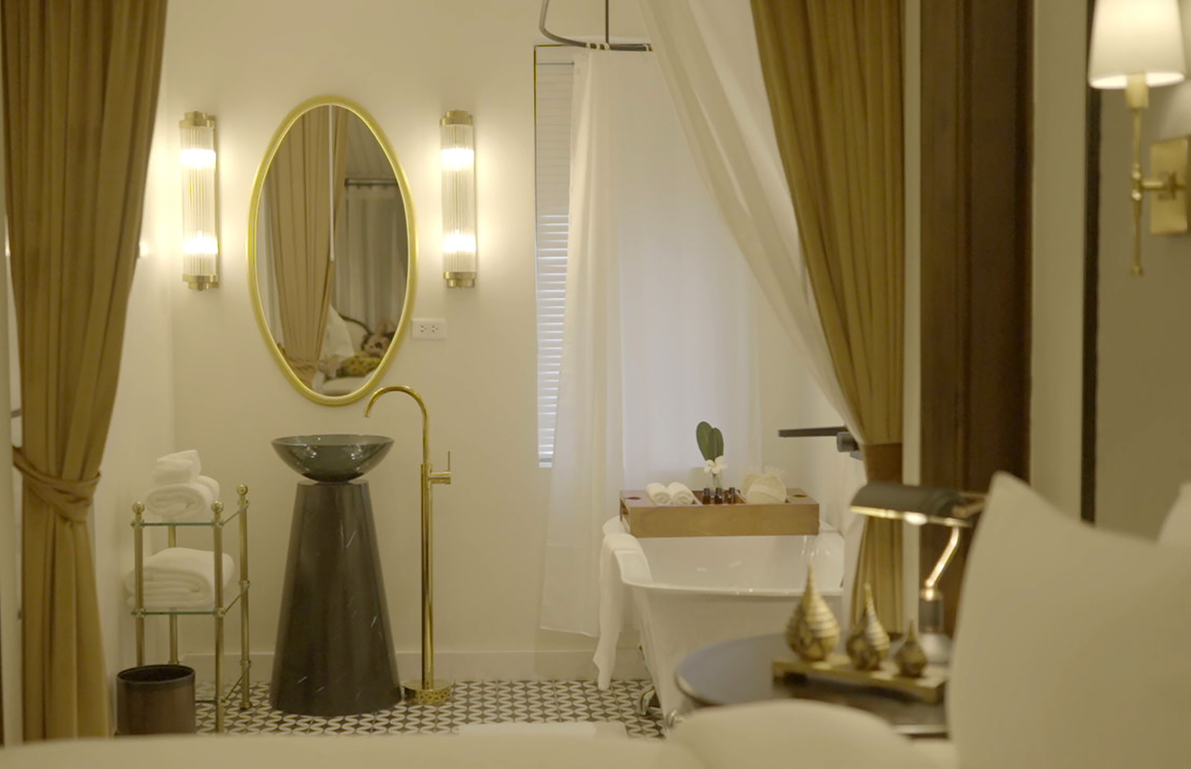 A bathroom with a large oval mirror, a sink with a bowl, a towel rack, and a vanity with a mirror and lights.