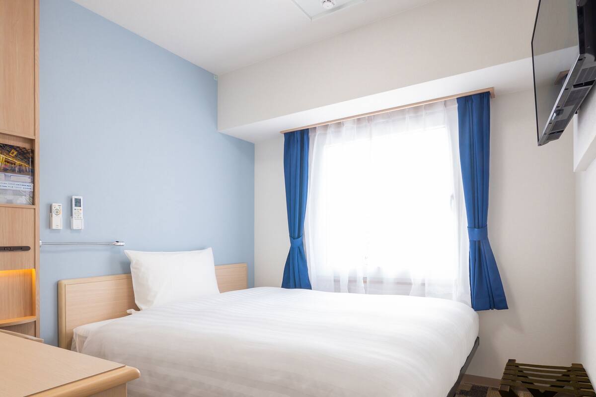 Una habitación de hotel con cama, escritorio, televisor y ventana con cortinas.