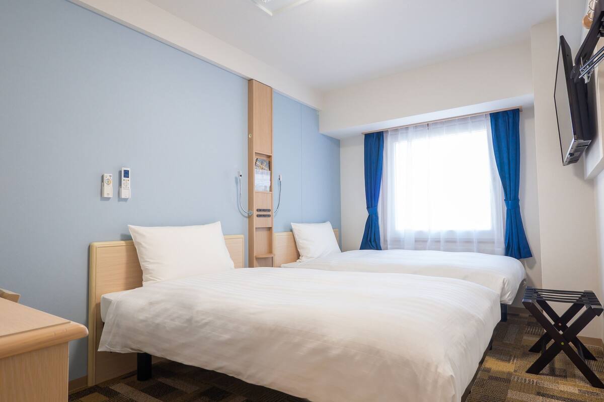 Habitación de hotel con dos camas, un armario de madera, una mesita de noche, un televisor montado en la pared y una ventana con cortinas azules.