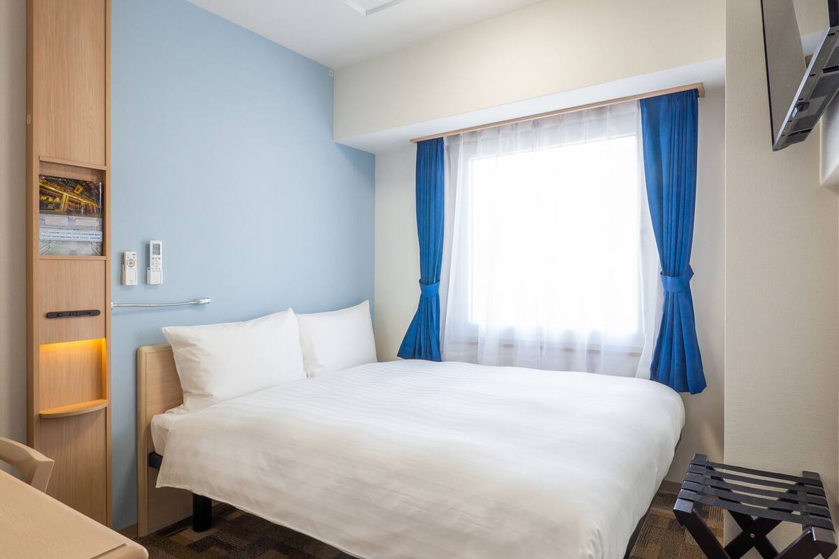 Una habitación de hotel con cama, escritorio, armario y ventana con cortinas.