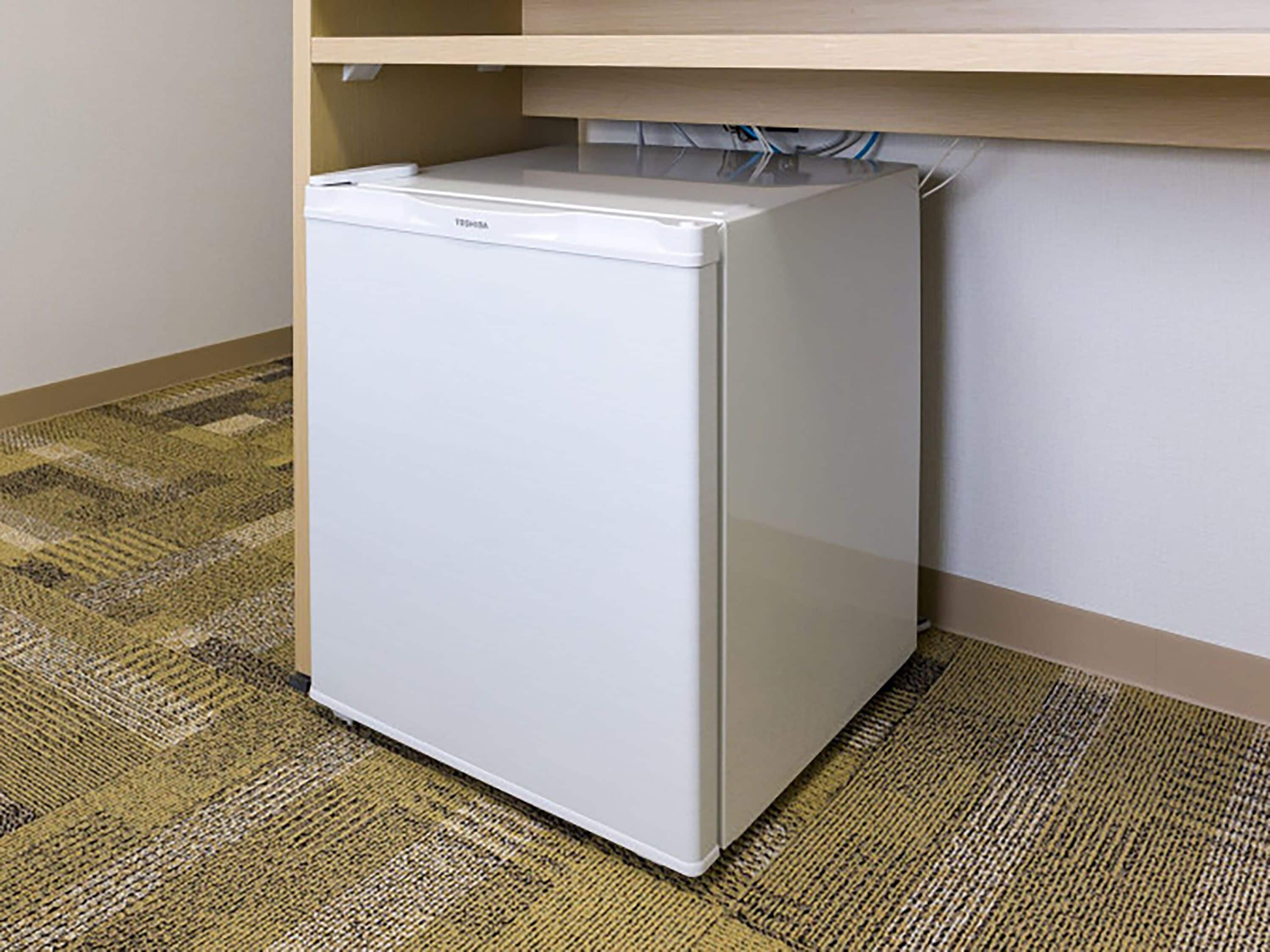 A compact white refrigerator in a corner of a room with a patterned carpet and a wooden shelf above it.