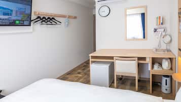 A hotel room with a bed, a desk with a chair, a small refrigerator, a wall-mounted TV, a clock, a mirror, a shelf with toiletries, and a hanging coat rack.