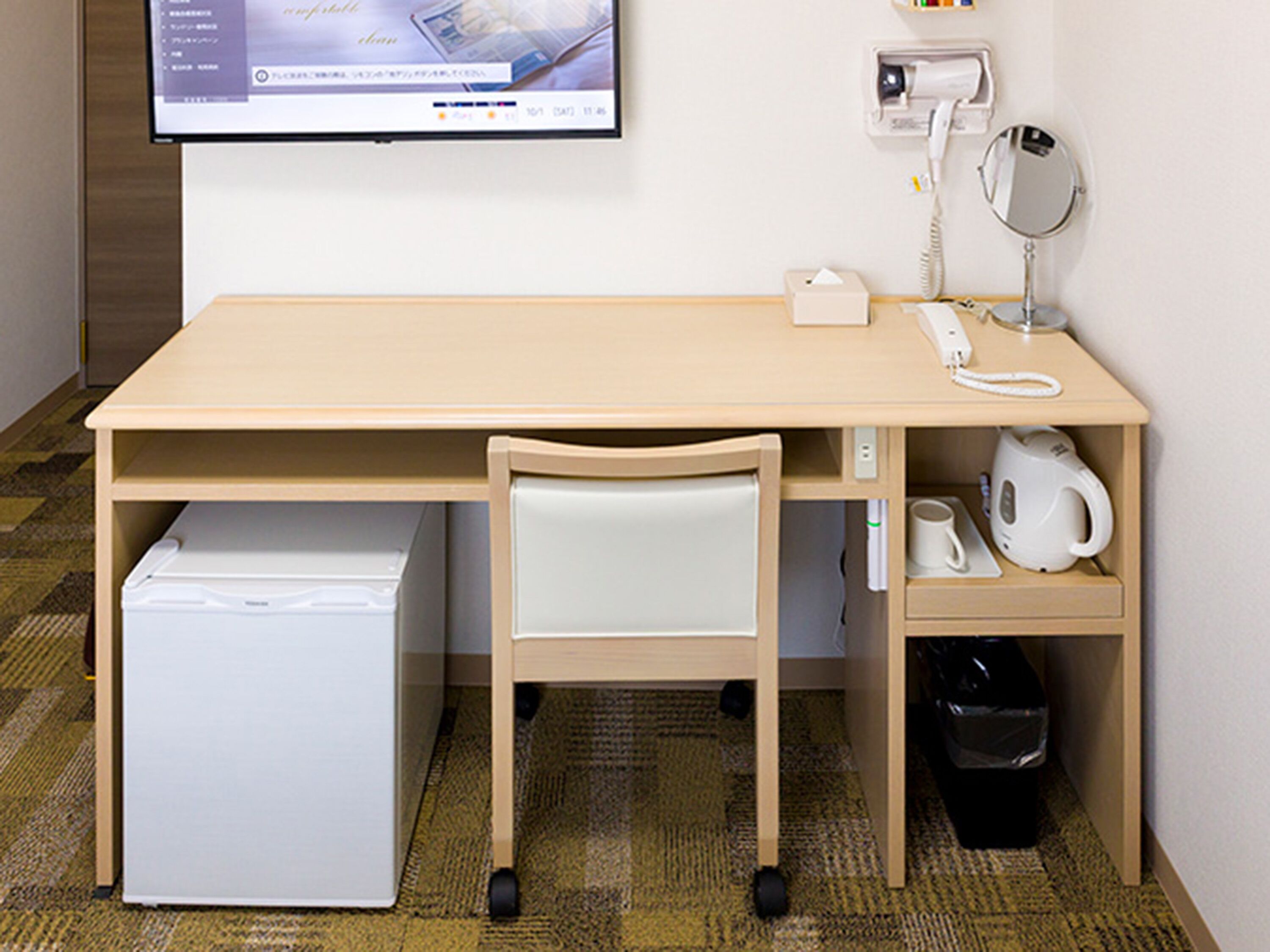 A compact hotel room with a wooden desk, a chair, a small refrigerator, a telephone, a mirror, a kettle, and a trash bin.