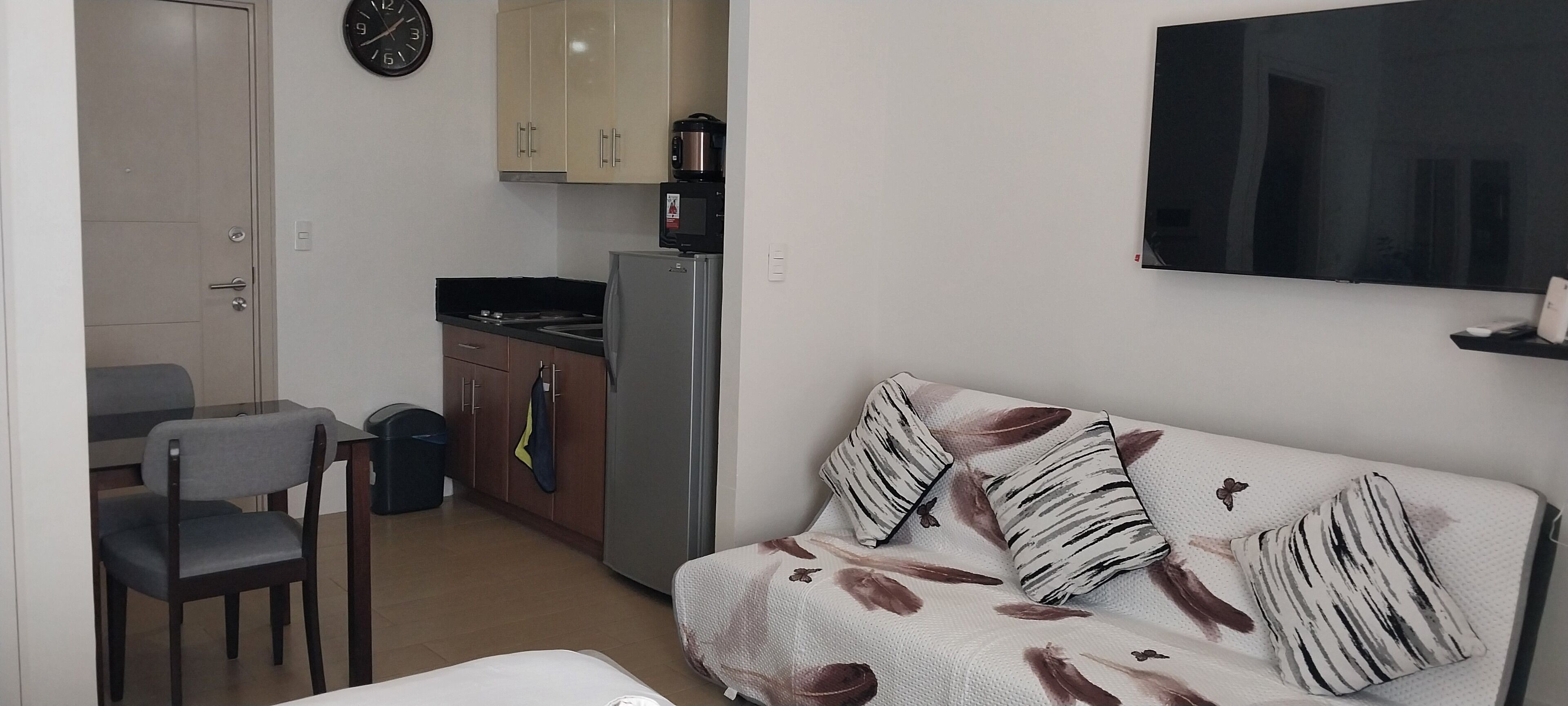 Un espace de vie compact comprenant un coin cuisine, un canapé agrémenté de coussins à motifs, un coin repas avec une chaise et un téléviseur fixé au mur.