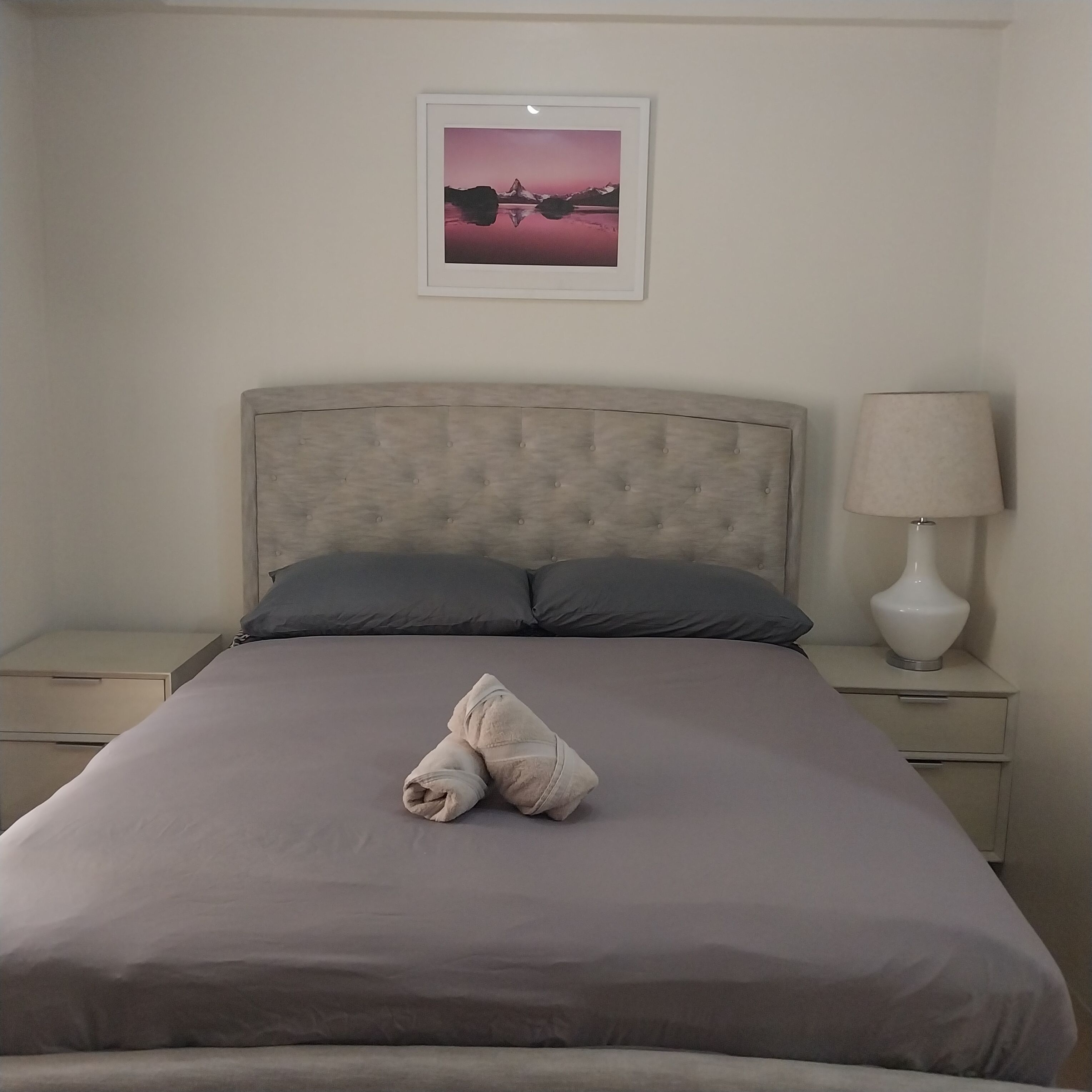 A neatly made bed with a grey headboard, a grey pillow, and a folded towel on the bed. There is a bedside table with a lamp on it. A framed picture hangs on the wall above the bed.